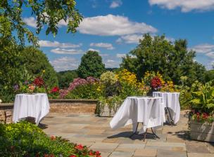 Three high top tables with white table cloths blowing in the wind on a patio surrounded by flowers.